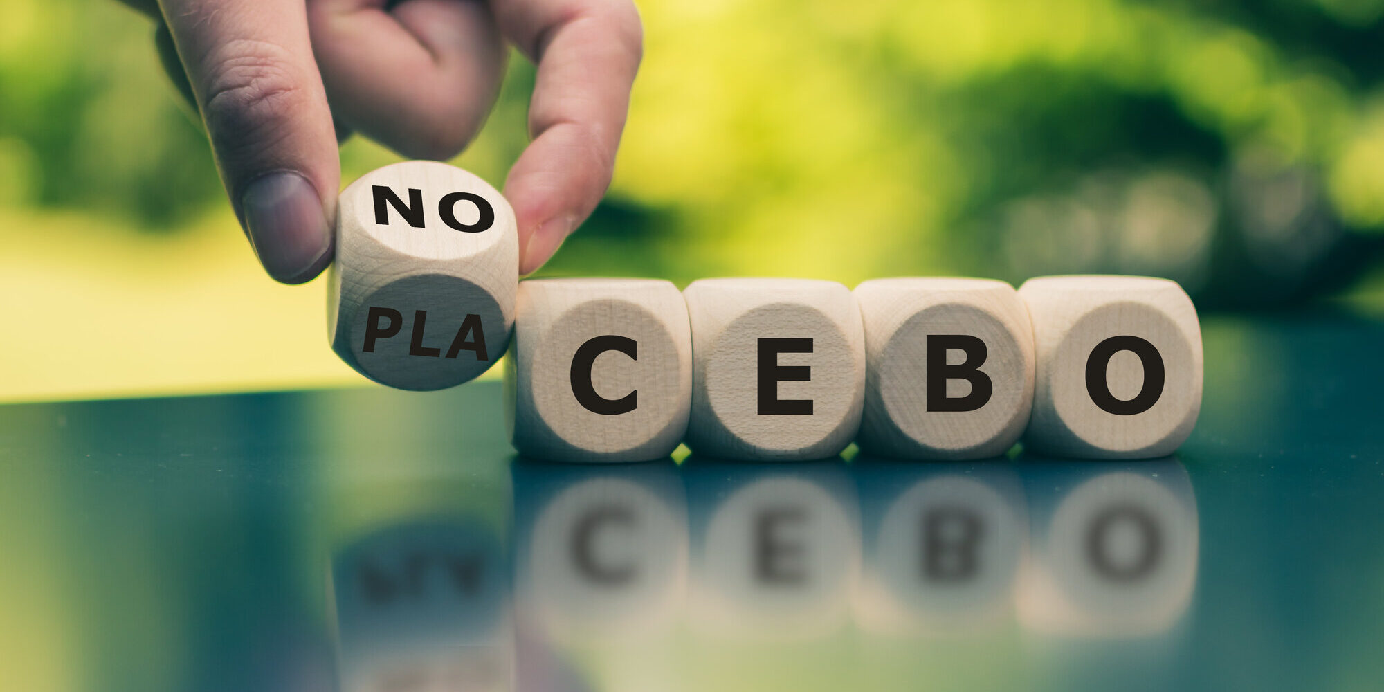placebo-nocebo.jpeg Holzwürfel mit Buchstaben, die das Wort 'NO PLACEBO' bilden, auf einem Tisch in der Natur.