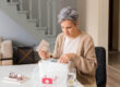 Eine Frau in einem hellen Raum untersucht einen durchsichtigen Medikamentenbehälter und hält eine Packung Tabletten.