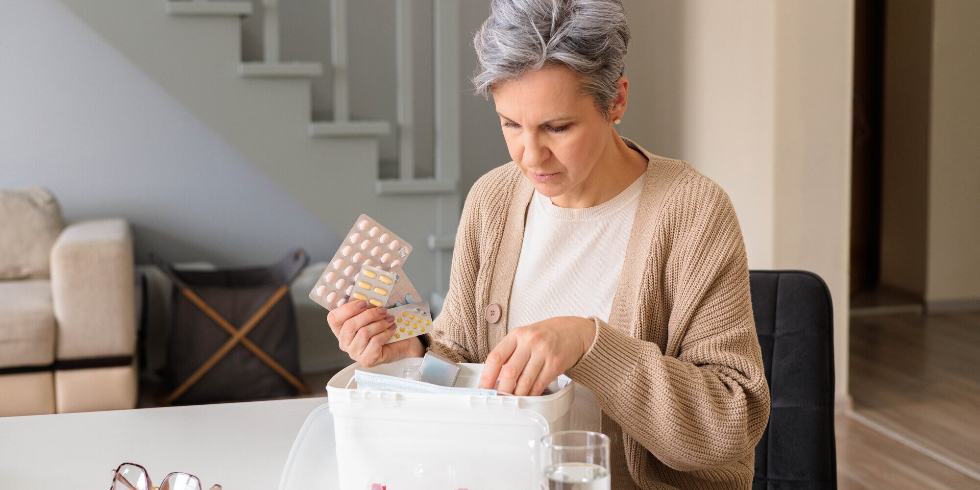 medikamente-lagerung.jpeg Eine Frau in einem hellen Raum untersucht einen durchsichtigen Medikamentenbehälter und hält eine Packung Tabletten.