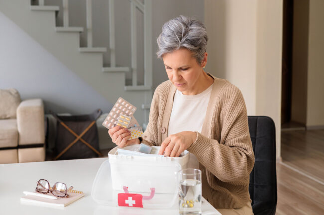medikamente-lagerung.jpeg Eine Frau in einem hellen Raum untersucht einen durchsichtigen Medikamentenbehälter und hält eine Packung Tabletten.